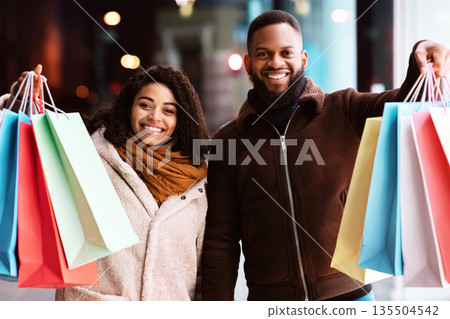 Sale And Consumerism Concept. Portrait of happy african american couple holding shopping bags, standing outdoors in the evening. Smiling black man and woman showing purchases outside the mall Sale And Consumerism Concept. Portrait of happy african american couple holding shopping bags, standing outdoors in the evening. Smiling black man and woman showing purchases outside the mall 135504542