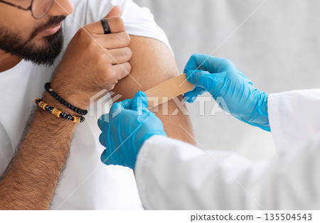 Cropped of arab man getting shot injection against coronavirus, closeup, copy space. Unrecognizable doctor in protective medical suit putting band on male patient shoulder, vaccination at home concept 135504543