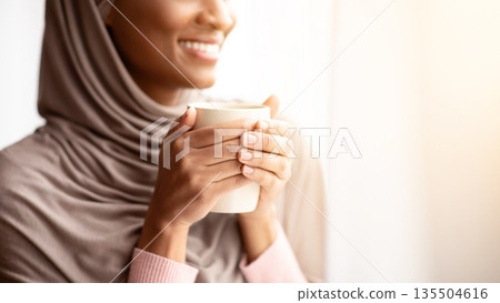 Cropped view of happy black woman in hijab warming her hands on cup of hot coffee near window at home. Lovely young Muslim lady having relaxing morning with aromatic drink, enjoying her break Cropped view of happy black woman in hijab warming her hands on cup of hot coffee near window at home. Lovely young Muslim lady having relaxing morning with aromatic drink, enjoying her break 135504616