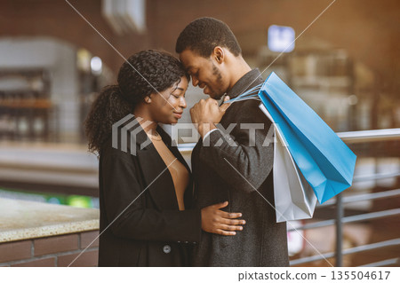 Romance for two, tenderness and date during seasonal sales and Black Friday. Young african american couple in love with colored shopping bags hugging at city mall ready to buy, side view, free space 135504617