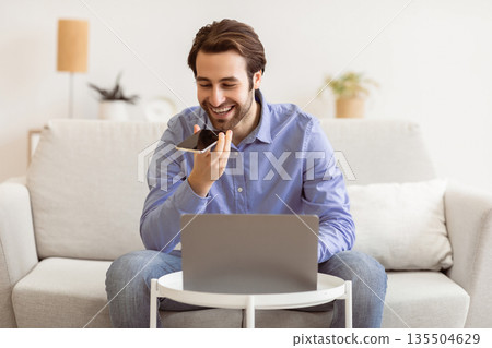 Smiling Businessman Talking By Phone In Loudspeaker Mode Or Using Voice Assistant On Smartphone Sitting At Laptop Computer On Couch Indoors. Gadgets And Modern Lifestyle, Voice Search Application 135504629