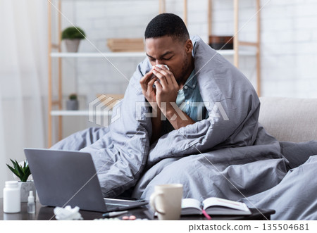 Ill African Man Having Rhinitis Sneezing And Blowing Runny Nose In Paper Tissue Sitting On Sofa At Home, Working Distantly On Laptop. Sinusitis Illness, Cold And Influenza Symptoms. Ill African Man Having Rhinitis Sneezing And Blowing Runny Nose In Paper Tissue Sitting On Sofa At Home, Working Distantly On Laptop. Sinusitis Illness, Cold And Influenza Symptoms. 135504681