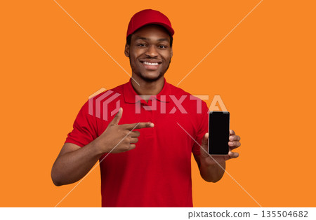 Choose Our Delivery Service. Smiling african american courier holding mobile phone with blank screen for mockup, wearing red cap and uniform, pointing at black empty gadget, orange studio background 135504682