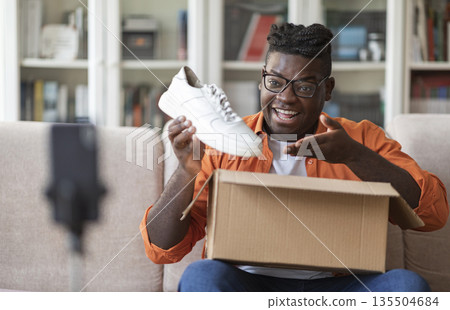 Social media influencer stylish cheerful chubby black guy reviewing product. Excited african american millennial man vlogging about unpacking white sneakers and filming himself at home on cell phone 135504684