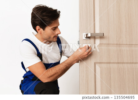 Repair Service Concept. Portrait of smiling handsome young locksmith in blue uniform installing door knob on front wooden entrance door at home or office workplace, isolated on white studio background Repair Service Concept. Portrait of smiling handsome young locksmith in blue uniform installing door knob on front wooden entrance door at home or office workplace, isolated on white studio background 135504695
