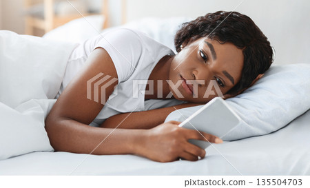 Frustrated young black woman looking at smartphone screen while laying in bed before sleeping, feeling lonely or depressed, closeup. Upset african american lady waiting for call from lover Frustrated young black woman looking at smartphone screen while laying in bed before sleeping, feeling lonely or depressed, closeup. Upset african american lady waiting for call from lover 135504703