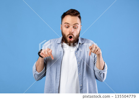 Shocked young bearded man pointing downward at blank space over blue studio background, free space. Amazed millennial man having amazing offer, offering big discount or sale 135504779