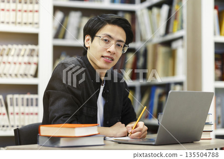 Working On Research Project. Portrait of asian male student using laptop at library, writing in notepad, looking at camera Working On Research Project. Portrait of asian male student using laptop at library, writing in notepad, looking at camera 135504788