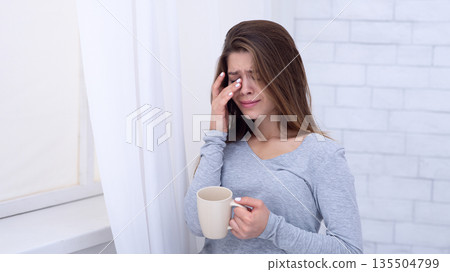Sad young woman with cup of coffee crying near window, wiping her tears at home. Depressed millennial lady feeling lonely, stressed or desperate, suffering from mental disorder 135504799