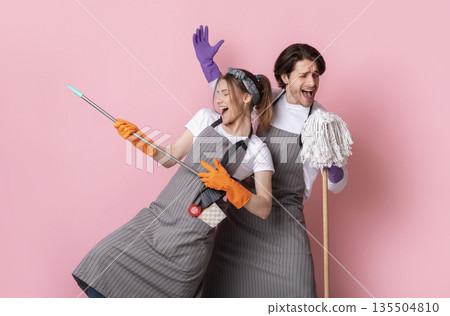 Playful couple of cleaners fooling together while making spring tidying, singing into mop and using broom as guitar over pink background 135504810