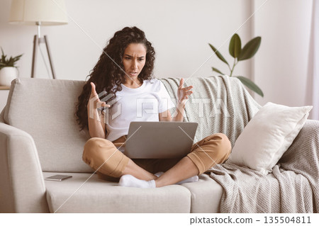 Angry woman freelancer looking at laptop screen and gesturing, living room interior, copy space. Emotional young brunette lady having problems with her job, sitting on couch at home Angry woman freelancer looking at laptop screen and gesturing, living room interior, copy space. Emotional young brunette lady having problems with her job, sitting on couch at home 135504811