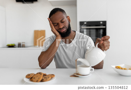 Lack Of Sleep Concept. Portrait of tired young African American man sleeping while sitting at dining table in kitchen, holding kettle pouring coffee away from cup spilling hot drink on desk Lack Of Sleep Concept. Portrait of tired young African American man sleeping while sitting at dining table in kitchen, holding kettle pouring coffee away from cup spilling hot drink on desk 135504812