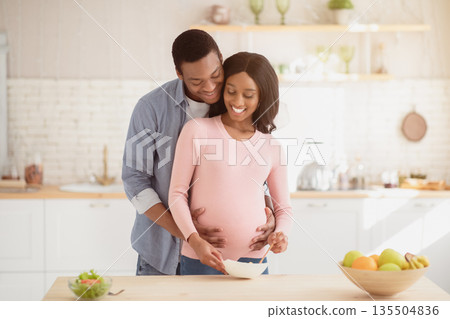 Lovely black pregnant woman making healthy breakfast in kitchen and husband helping her. African American expectant couple cooking balanced meal, spending time together indoors Lovely black pregnant woman making healthy breakfast in kitchen and husband helping her. African American expectant couple cooking balanced meal, spending time together indoors 135504836
