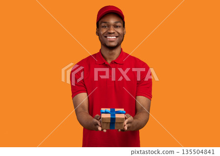 Delivery Service For Holidays. Portrait of smiling cheerful african american courier guy holding wrapped gift box, giving and showing present to camera, isolated over orange studio background 135504841
