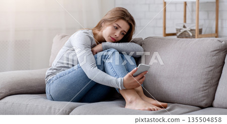 Portrait of depressed young woman sitting on couch with smartphone in her hands. Millennial lady suffering from cyber bullying or going through bad breakup. Mental health concept Portrait of depressed young woman sitting on couch with smartphone in her hands. Millennial lady suffering from cyber bullying or going through bad breakup. Mental health concept 135504858