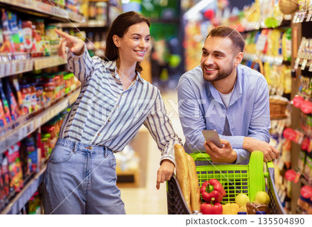 I Want This. Millennial Couple Doing Grocery Shopping Together, Man Leaning On Cart And Using Mobile Phone, Woman Pointing At Shelf, Family Choosing Food Products In Supermarket And Buying Supplies I Want This. Millennial Couple Doing Grocery Shopping Together, Man Leaning On Cart And Using Mobile Phone, Woman Pointing At Shelf, Family Choosing Food Products In Supermarket And Buying Supplies 135504890