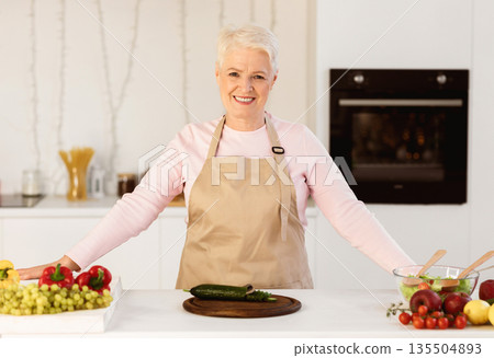 Happy Senior Lady Cooking In Kitchen Making Salad For Dinner At Home, Smiling To Camera. Aged Woman Preparing Healthy Meal Posing Indoors. Nutrition And Recipes For Older Female Happy Senior Lady Cooking In Kitchen Making Salad For Dinner At Home, Smiling To Camera. Aged Woman Preparing Healthy Meal Posing Indoors. Nutrition And Recipes For Older Female 135504893