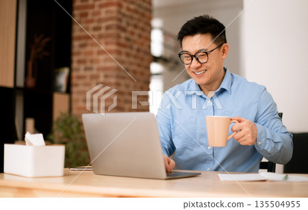 Asian middle aged man drinking coffee at home office, sitting at workplace with computer, free space. Business, coffee break from work, pause and vitality for freelance Asian middle aged man drinking coffee at home office, sitting at workplace with computer, free space. Business, coffee break from work, pause and vitality for freelance 135504955