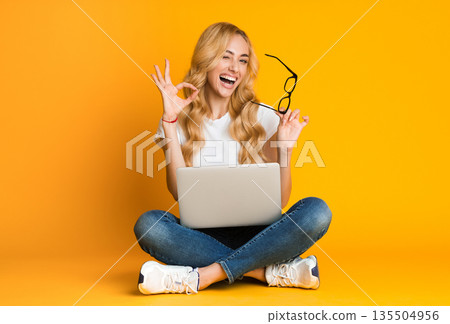 It's okay. Overjoyed woman showing ok sign, sitting with laptop over yellow background 135504956