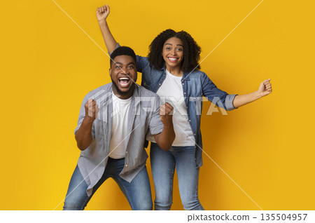 Lucky Winners. Portrait Of Euphoric Young Black Couple Emotionally Celebrating Success, Raising Fists And Shouting In Excitement On Yellow Background 135504957