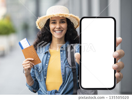 Happy attractive millennial brunette lady in straw hat tourist holding passport with flight tickets and cell phone with white blank screen, showing nice mobile app for travellers, mockup 135505020