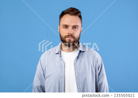 Portrait of serious young guy looking at camera on blue studio background. Successful millennial man in shirt with dark hair and beard posing, feeling confident and focused Portrait of serious young guy looking at camera on blue studio background. Successful millennial man in shirt with dark hair and beard posing, feeling confident and focused 135505021