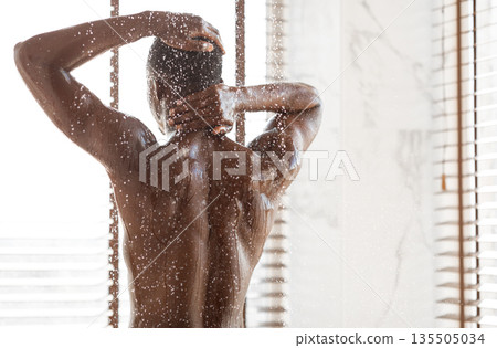 Unrecognizable African American Man Taking Shower Washing Body Standing Back To Camera Under Falling Water Drops In Modern Bathroom Indoor. Male Bodycare, Wellness And Spa. Back View, Copy Space 135505034