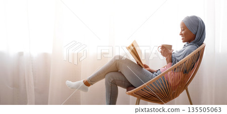 Side view of young black woman in hijab drinking coffee and reading in cozy armchair indoors, copy space. Millennial Islamic woman in traditional headscarf enjoying hot beverage and book at home Side view of young black woman in hijab drinking coffee and reading in cozy armchair indoors, copy space. Millennial Islamic woman in traditional headscarf enjoying hot beverage and book at home 135505063