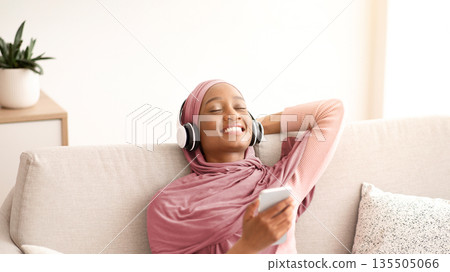 Positive young black woman in hijab relaxing on sofa with headphones, listening to music or audio book at home. Lovely Muslim lady in traditional headscarf enjoying beautiful song on smartphone 135505066
