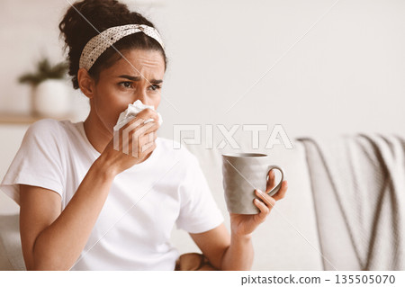 Young woman feeling sick, sneezing her nose, holding mug with hot drink and napkin, home interior, looking at copy space. Ill brunette lady having runny nose, got cold or coronavirus infection 135505070