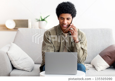 E-learning concept. Funky black male student communicating on cellphone in front of laptop computer at home. Handsome teenage guy participating in online class or web conference indoors 135505090