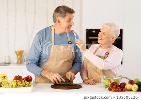 Aged Spouses Cooking And Tasting Food Feeding Each Other Having Fun During Dinner Preparation Standing In Modern Kitchen At Home. Senior Family Couple Enjoying Weekend Preparing Meal Together. 135505120