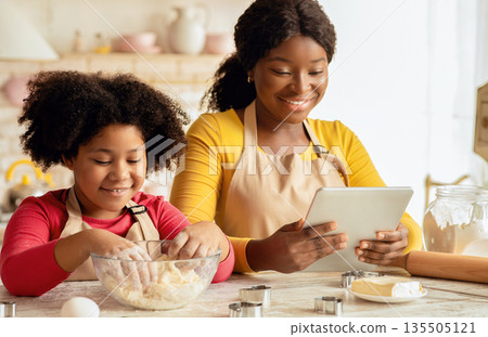 Cheerful Black Mom And Cute Little Child Using Digital Tablet In Kitchen While Baking, Checking Cookies Recipe Online, African American Family Enjoying Preparing Homemade Pastry, Closeup Shot 135505121