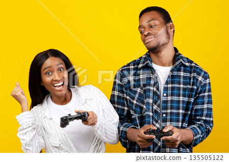Winning and losing in online games at home and human emotions. Young sad african american guy looking at happy wife with gamepad in hands, isolated on yellow background, studio shot, free space 135505122