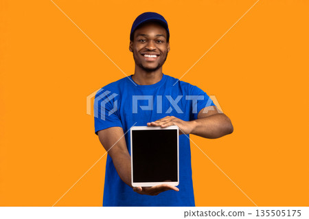 Delivery Application. African american courier man holding digital tablet with blank screen for mock up and showing it to camera, wearing blue cap and uniform, standing isolated on orange background Delivery Application. African american courier man holding digital tablet with blank screen for mock up and showing it to camera, wearing blue cap and uniform, standing isolated on orange background 135505175