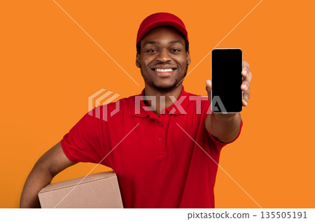 Delivery Service Application. Smiling african american man showing smartphone with blank screen for mockup, wearing red cap, uniform and thermo backpack bag, holding box, isolated on orange background Delivery Service Application. Smiling african american man showing smartphone with blank screen for mockup, wearing red cap, uniform and thermo backpack bag, holding box, isolated on orange background 135505191
