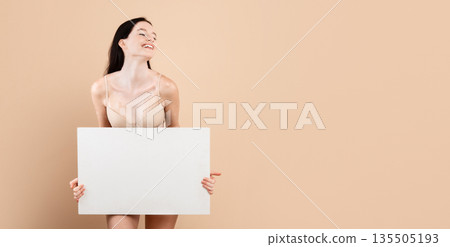 Smiling young female with vitiligo skin disorder holding white blank placard, beautiful millennial lady standing in underwear over beige background in studio, enjoying her different beauty, copy space 135505193