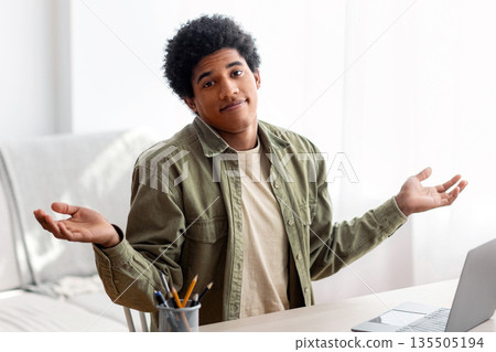 Puzzled black student sitting in front of laptop, not understanding difficult lesson, having difficulty studying online. African American teen guy shrugging shoulders, feeling confused during exam Puzzled black student sitting in front of laptop, not understanding difficult lesson, having difficulty studying online. African American teen guy shrugging shoulders, feeling confused during exam 135505194