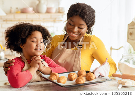 Happy little black girl looking at tray with freshly baked croissants while cooking with mom in kitchen, adorable female child and her mother wearing aprons enjoying preparing homemade food, closeup 135505218