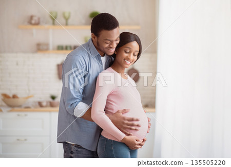 Pregnant black woman and her happy husband hugging next to window at home. Handsome African American man touching his expectant wife's tummy, spending cozy moment together as family Pregnant black woman and her happy husband hugging next to window at home. Handsome African American man touching his expectant wife's tummy, spending cozy moment together as family 135505220