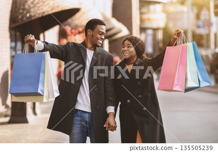 Best price for Black Friday and season sale. Cheerful millennial african american male and female look at each other, shows many colored shopping bags, expressed success, near city mall, outdoor 135505239