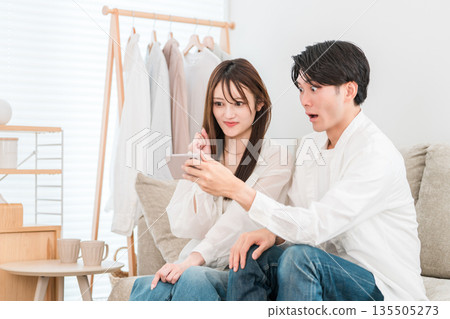 一對年輕的亞洲夫婦坐在家裡的客廳沙發上，看著他們的智慧型手機。 135505273