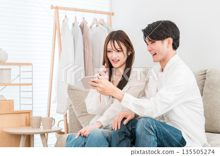 一對年輕的亞洲夫婦坐在家裡的客廳沙發上，看著他們的智慧型手機。 135505274