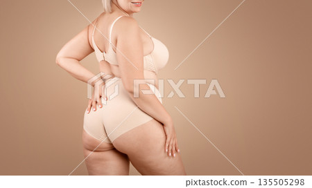 Young plus size woman in beige underwear posing over brown studio background, half-turned at camera, cropped image with free space 135505298