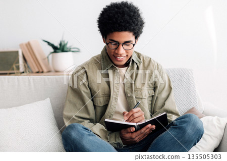 Focused African American student taking notes in copybook, working on school project at home. Black male teenager learning remotely, making homework, getting ready for online lesson Focused African American student taking notes in copybook, working on school project at home. Black male teenager learning remotely, making homework, getting ready for online lesson 135505303