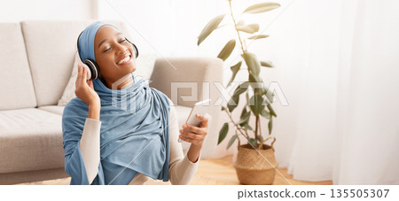 Full length portrait of pretty Muslim lady in hijab and headset listening to music on floor at home. African American woman in Arab scarf enjoying interesting audio book or nice melody Full length portrait of pretty Muslim lady in hijab and headset listening to music on floor at home. African American woman in Arab scarf enjoying interesting audio book or nice melody 135505307