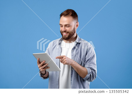 Joyful bearded man using tablet computer for online work or studies on blue studio background. Positive teacher giving remote lesson. Blogger creating new post for social network Joyful bearded man using tablet computer for online work or studies on blue studio background. Positive teacher giving remote lesson. Blogger creating new post for social network 135505342