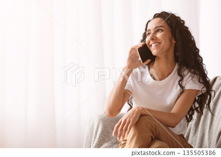 Happy young brunette lady with long curly hair in casual sitting by window at home, cheerfully talking on mobile phone with friends or lover, copy space. Communication during isolation concept Happy young brunette lady with long curly hair in casual sitting by window at home, cheerfully talking on mobile phone with friends or lover, copy space. Communication during isolation concept 135505386