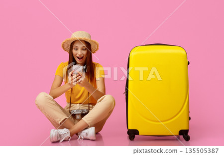 Amazed young woman sitting by suitcase and using mobile phone over pink studio background, found great deals for travelling. Active resting, vacation, tourism, travelling concept Amazed young woman sitting by suitcase and using mobile phone over pink studio background, found great deals for travelling. Active resting, vacation, tourism, travelling concept 135505387