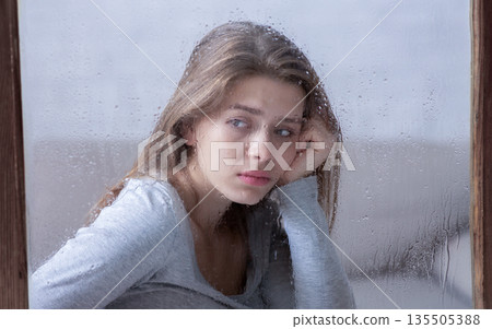 Sad young woman sitting at home and looking out window at falling rain. Lonely lady suffering from autumn depression, feeling alone, hopeless or melancholic. Mental health concept Sad young woman sitting at home and looking out window at falling rain. Lonely lady suffering from autumn depression, feeling alone, hopeless or melancholic. Mental health concept 135505388